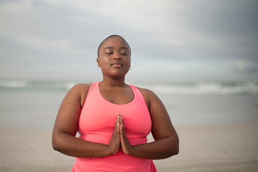 Meditation, prayer hands and plus size black woman at a beach for peace, zen or mental health wellness. Mindfulness, healing or African lady person at sea for gratitude, balance or holistic breathing
