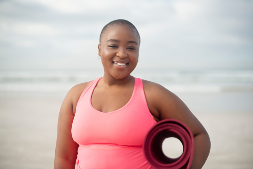 Yoga mat, portrait and plus side black woman at a beach for meditation, wellness and stress relief in nature. Face, smile and happy African female person at sea for holistic mental health exercise