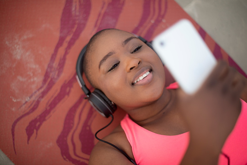 Woman, selfie and happy with headphones for music with relax, social media update or podcast on beach. Black person, smartphone and lying down outdoor with radio, streaming audio or video call