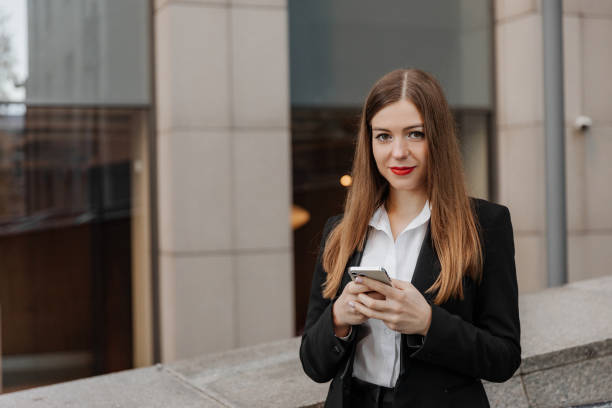 istock Young business woman wearing black dress code smiling answering message or chatting, using phone and looking at the camera 1959843915