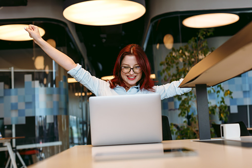 A beautiful young woman, who could be a design worker, trainee, or businesswoman, exudes joy and celebration in an open, modern office work area. With her arms extended outwards, she signifies a moment of triumph, possibly after a successful business deal.
Business deal, beautiful, young woman, design worker, trainee, businesswoman