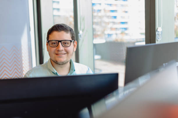 istock Portrait of a smiling young IT specialist or computer software programmer happy male with reading eyeglasses looking at camera while working at his workplace and typing codes on a computer behind of lots of monitors 1959853434
