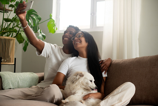 Happy couple, dog and selfie with hug, home and living room with embrace, love, and couch. Boyfriend, girlfriend and smile for social media, bonding together and intimate romantic relationship