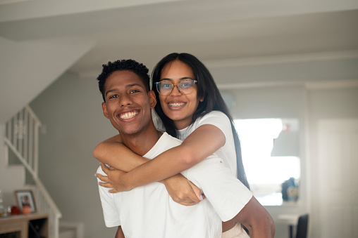 Couple, portrait and smile in home with piggyback for bonding, relationship and relax in living room. Young people, face and happy with embrace or fun for love, care and playful in apartment or house