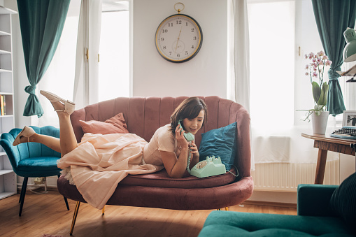 Classy young woman talking on wired landline vintage telephone while lying on sofa in her modern apartment.