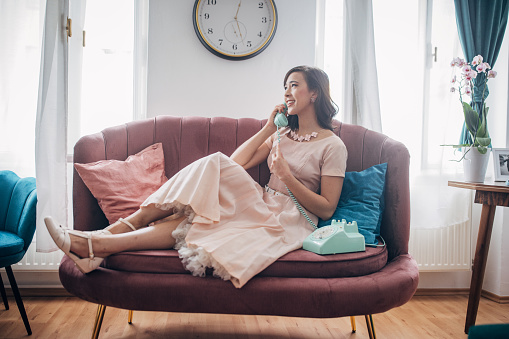 Classy young woman talking on wired landline vintage telephone while sitting on sofa in her modern apartment.