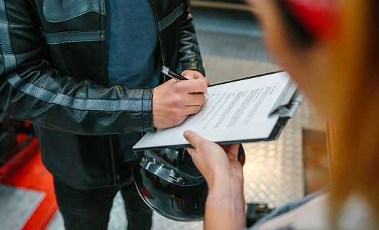 Close up of unrecognizable biker man wearing leather jacket and holding helmet signing insurance policy to receipt his repaired motorcycle on workshop while female mechanic holding the clipboard