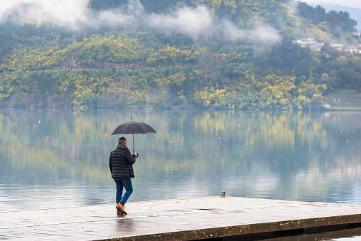 Castrelo de Miño, Spain - February 12, 2017: A man with an umbrella, on a jetty, observes a reservoir where the landscape is reflected.