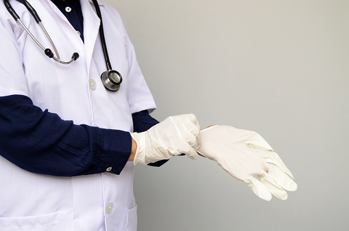 Horizontal photograph of one unidentifiable surgeon with blue shirt, white lab coat, stethoscope getting ready by putting on surgical gloves in her hand over grey background with copy space for text. The person is at the left side leaving right side free for text. Just the body is visible while the face is not.