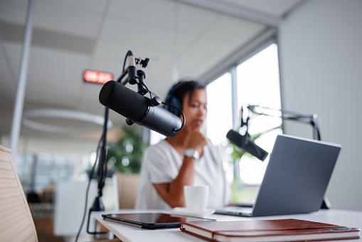 A Black mid adult woman thoughtfully adjusts her podcast microphone, ensuring optimal sound quality before going live, with her laptop ready for the session.