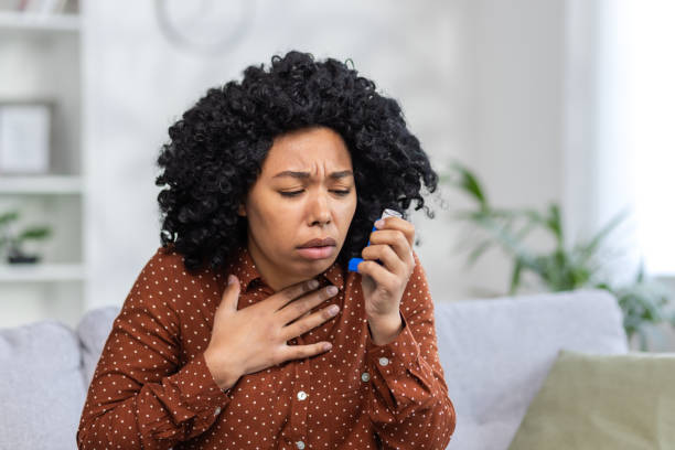 foto ravvicinata di una donna afroamericana seduta sul divano di casa, che non si sente bene, respira a fatica, si tiene il petto e usa uno spray inalatore - torso physical therapy patient relaxation exercise foto e immagini stock