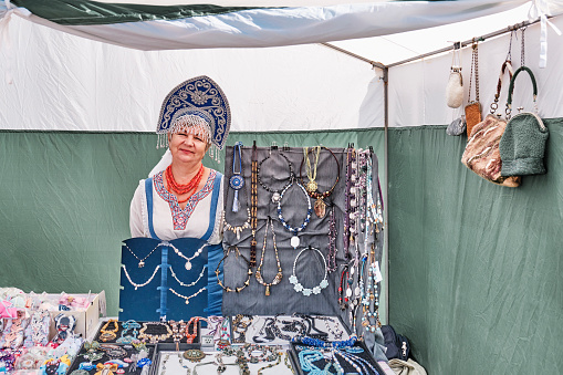 Irbit, Russia - August 11, 2023: Senior Caucasian smiling saleswoman in Russian national clothes and embroidered kokoshnik. Selling costume jewelry and accessories in trade tent at traditional fair.