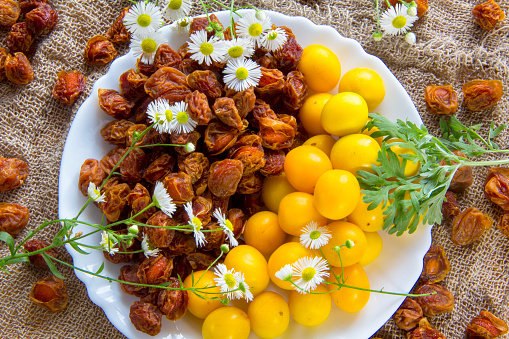 Fresh yellow plum berries and Dried orange plum berrieson a white plate. White daisy flowers and a green wormwood branch. Healthy food. Edible still life. Berries and flowers of chamomile. Dry fruits for compote, health.