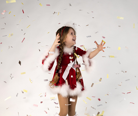 Happy Caucasian little girl wearing Christmas themed dress on gray studio background, copy space. Portrait of beautiful child having fun with sparkling confetti. High quality photo