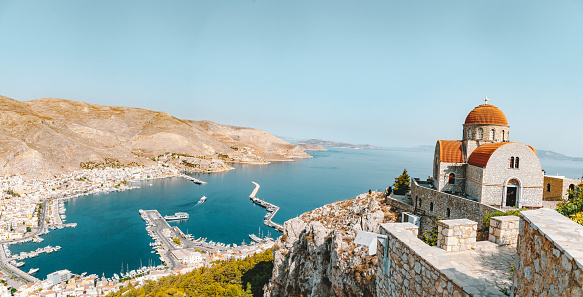 A breathtaking aerial view captures the beauty of Kalymnos island in the Dodecanese, Greece. Bathed in sunlight, Pothia town unfolds along the coastline, while the picturesque Agios Savvas Monastery stands against the vivid blue sky and the mesmerizing expanse of the Aegean Sea. A perfect snapshot of the island's charm on a tranquil summer day.