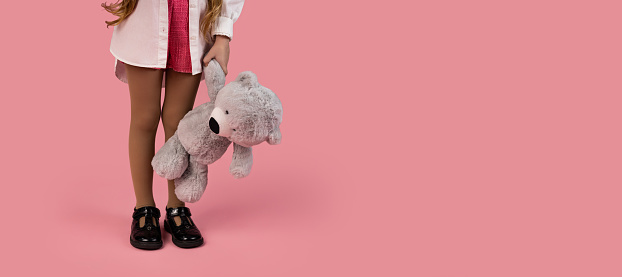 A little girl is holding a gray plush toy, more precisely a teddy bear on a pink background, the little girl is very stylishly dressed, ready for Valentine's Day. High quality photo