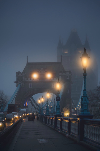 London's iconic Tower Bridge and the majestic Thames River come alive in the ethereal embrace of a winter dawn fog. The soft glow of street lamps illuminates the atmospheric scene, while cars cautiously traverse the misty streets, creating a captivating and serene winter morning in the heart of the United Kingdom capital.