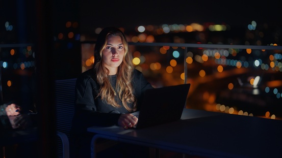 A portrait of a young woman using her laptop at balcony at home at night.