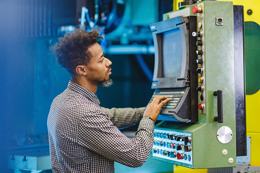 A mixed adult male electrical engineer setting up a CNC machine in a production facility. He is adjusting the final settings. The engineer is pressing the keys on the keyboard while checking the adjustments on the screen. He is focused.