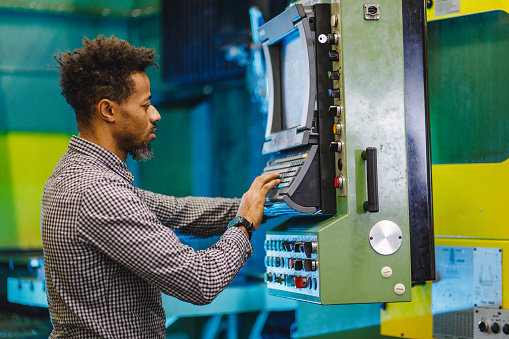 A mixed adult male production manager controlling the CNC machine. He is standing by a control panel of the machine and touching the keys on the keyboard. He looks engrossed in work and serious. He is located in a production facility