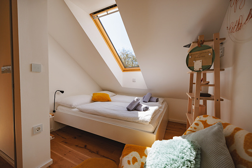 A cozy and bright bedroom at the attic of a house has a bed with white sheets and some towels ready to be used for the guests. In front there is a sofa with some cushions and next to it a wooden bookshelf with some things.