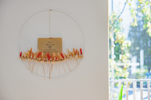 A colorful ornament in orange and beige color with a message in the middle is hanging on the white wall inside an apartment. At the back there is a window where daylight is coming from the outside and some plants can be seen.