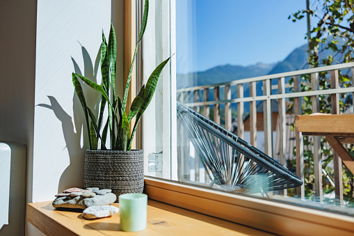 Big window with wooden edges decorated with a plant inside a flower pot and some stones and a candle next to it. Through the window there is a beautiful landscape with a mountain at the back and some chairs at a balcony.
