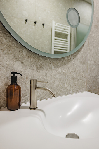 Close up of a basin with a water faucet located at a modern bathroom of an apartment. There is a circular mirror at the top which reflects the shower and the heater at  the back.