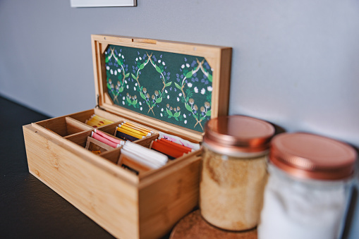 A nice wooden box with decoration inside is open and containing many colorful tea sachets. Next to it there are two glass containers with white and borwn sugar closed with a metallic cover.