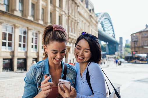 Two women wearing fashionable, casual clothing, walking through Newcastle City Centre, England. They are smiling as they look at a selfie they have taken on a smartphone.