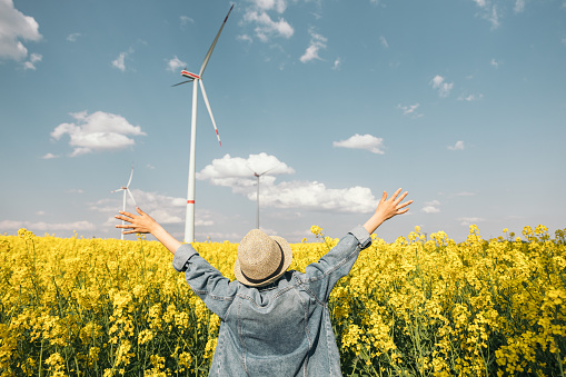 Teenage girl in hat and denim jacket with her back turned, opening her arms in a windmill field with yellow flowers