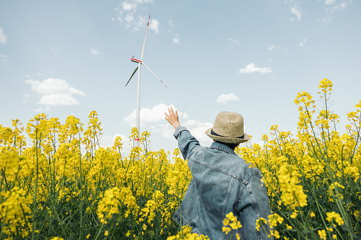 Teenage girl with her back turned with her hand up pointing at a wind turbine in a field of yellow flowers