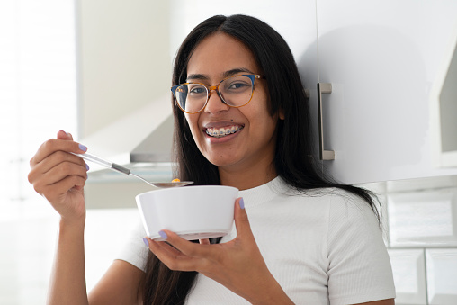 Happy woman, braces and eating porridge in bowl for morning, breakfast or meal in kitchen at home. Portrait of female person smile with glasses for delicious snack, fiber or calcium vitamins at house