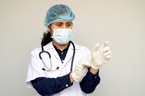 Horizontal photograph of one female surgeon with blue shirt, white lab coat, stethoscope getting ready by putting on surgical gloves in her hand over grey background with copy space for text.