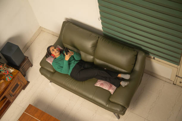 Directly above shot of cheerful young woman with smart phone listening music over headphones while lying on sofa in living room