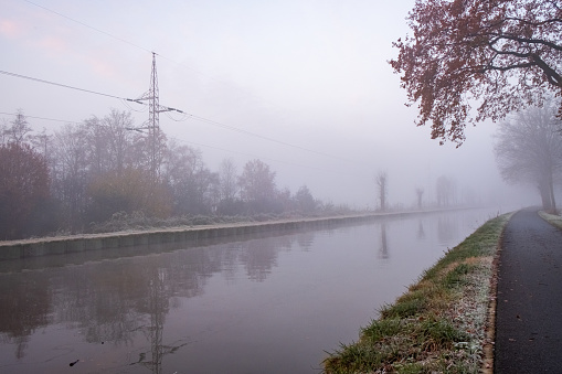 This photograph captures a tranquil early morning scene by a canal, veiled in a dense, soft fog. The mist shrouds the surrounding trees and power lines, creating a minimalist landscape that conveys solitude and silence. The water is still, reflecting the faint outlines of the trees and the grey sky above. The frosted grass on the bank and the autumnal leaves of the overhanging tree hint at the cold, crisp air of the season. This image portrays the still and quiet beauty of a foggy dawn in a subdued, pastel palette. Fog Enshrouded Canal at Dawn. High quality photo