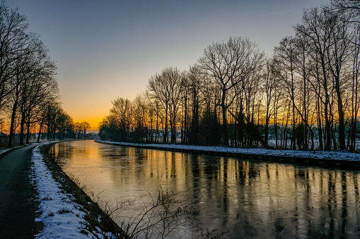 The image offers a serene view of a frozen canal lined by a snow-dusted path and skeletal trees during the twilight hours. As the sun dips below the horizon, its diminishing light casts a subtle array of colors in the sky, mirrored by the ice's glossy surface. The bare branches of the trees form a stark contrast against the sky, reflecting the day's end in this quiet natural setting. The peacefulness of the scene is palpable, inviting the viewer to savor the stillness of the winter evening. Dusk's Reflection on the Frozen Canal. High quality photo