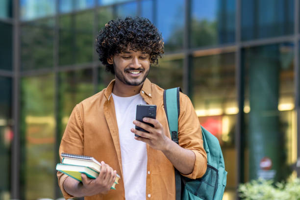 istock Smiling young Indian male student standing outside university campus with backpack and books and using mobile phone 1960207945