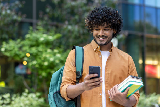 istock Indian smiling young male student standing outside with backpack and books and using mobile phone 1960208471