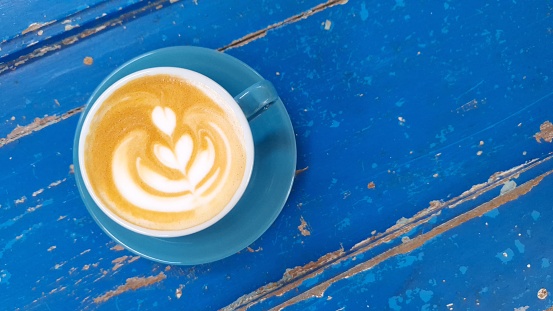 Top view of hot vanilla coffee latte and heart shaped latte art in blue glass with blue wooden table background and empty space for text on right side.