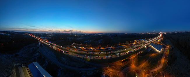 This panoramic image stretches across the E19 highway near Halle, leading towards Brussels, as the last light of day fades into the onset of evening. The highway is a band of activity with vehicles' lights creating a snaking pattern of illumination. The sky transitions from the warm tones of sunset on the right to the deepening blues of twilight on the left, symbolizing the day's end. The panoramic view encapsulates a vast landscape, with silhouettes of distant buildings against the sky and the glow of the city on the horizon. The photograph emphasizes the breadth of the transition from rural outskirts to the bustling urban center. Panoramic Dusk: E19 Near Halle with Brussels Horizon. High quality photo