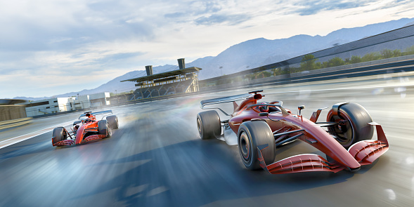 Two generic red and silver racing cars moving a high speed with motion blur. The race cars are racing past an empty grandstand in slightly wet conditions, emitting sprays from wheels, under a bright and cloudy sky. Location is fictional.