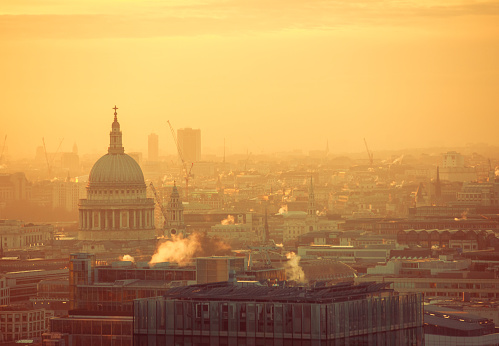 London's iconic St. Paul's Cathedral stands tall against the vibrant hues of a sunset-painted sky, casting a silhouette over the meandering River Thames. The cityscape is veiled in a delicate mist, and chimney smoke whispers tales of London's rich history. A breathtaking aerial view capturing the essence of a serene evening in the heart of the city.