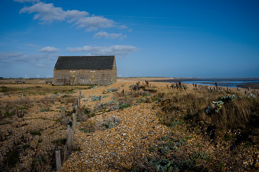 A shot of Rye Harbour including the Mary Stanford Boathouse and showing a shingle beach, groynes, a receding tide and a huge deep blue sky, with wind turbines on the horizon.