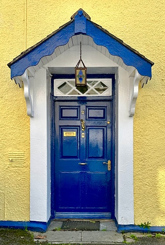 Bright blue, panel door with knocker and mail slot; white trim and peaked overhang with blue scalloped trim; hanging stained glass lantern.