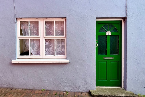 Single bright green door with panel windows and half moon upper window. White trimmed double sashes window with lace curtains on light blue stucco wall.