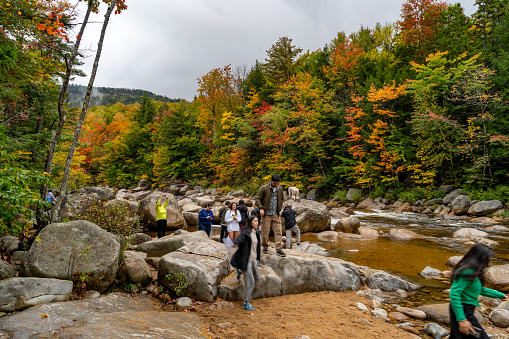 Swift River, New Hampshire, USA, October 2023. Autumnal leaf coloured forests on the Kancamagus Highway towards White Mountain.  This shows the Swift River running through the forest with tourist walking over the boulders at the side of the river.