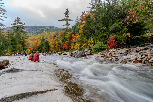 Swift River, New Hampshire, USA, October 2023. Autumnal leaf coloured forests on the Kancamagus Highway towards White Mountain.  This shows the Swift River running through the forest with tourist walking over the boulders at the side of the river.