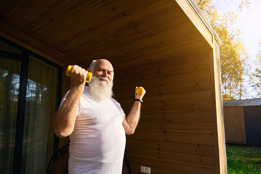 Cheerful old man does morning exercises with dumbbells, he exercises in the fresh air