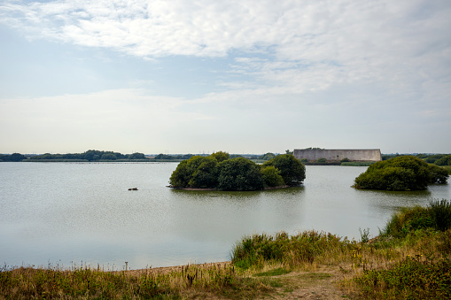 A wide shot of concrete sound mirrors on a calm and warm looking day, looking over a lake, in the Romney Marshes.